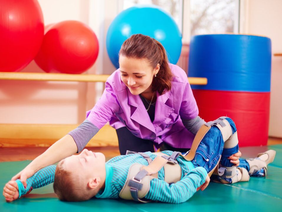 Hoitaja kuntouttaa vammaista lasta lattiapehmusteen päällä terapiatilassa. 
